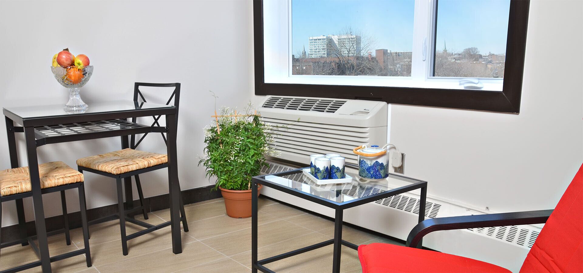 A room with a table, chairs, a potted plant, a window with a city view, and an air conditioning unit.