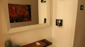 Un pasillo con un estante de madera que contiene un control remoto y adornos, un espejo enmarcado que refleja un cuadro, y una puerta con un detalle decorativo.