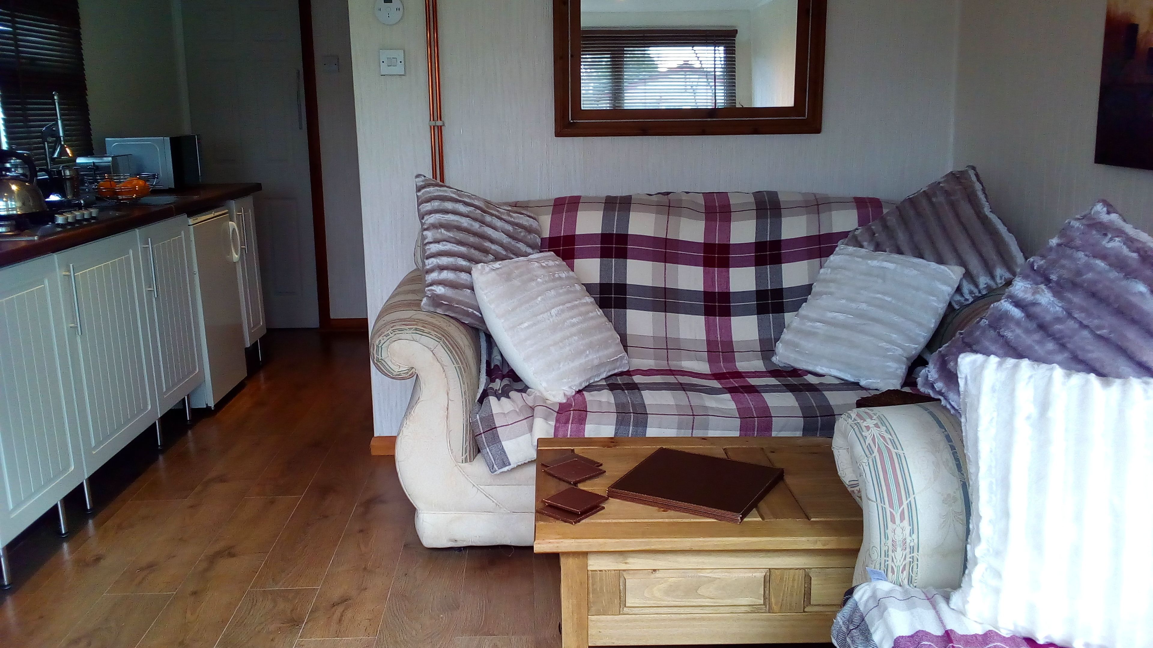 A cozy living room with a plaid sofa, a wooden coffee table, and a kitchen area with a kettle and oranges.