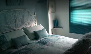 Quarto com cama, almofadas, mesa de cabeceira e janela com persianas.