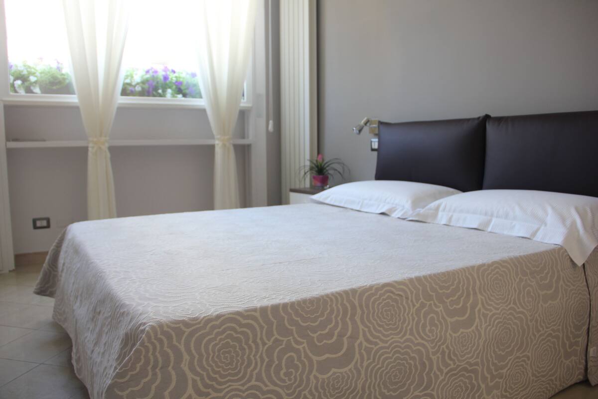 A neatly made bed with a patterned bedspread, a bedside table with a lamp, and a window with curtains.