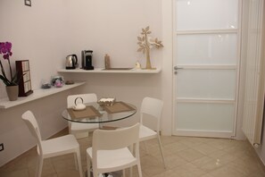 Un pequeño comedor con una mesa de vidrio y sillas blancas, una cafetera y un árbol decorativo en una estantería.