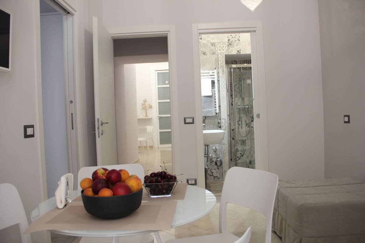 A dining area with a table set for two, a bowl of fruit, and a view into a bathroom with a shower and sink.