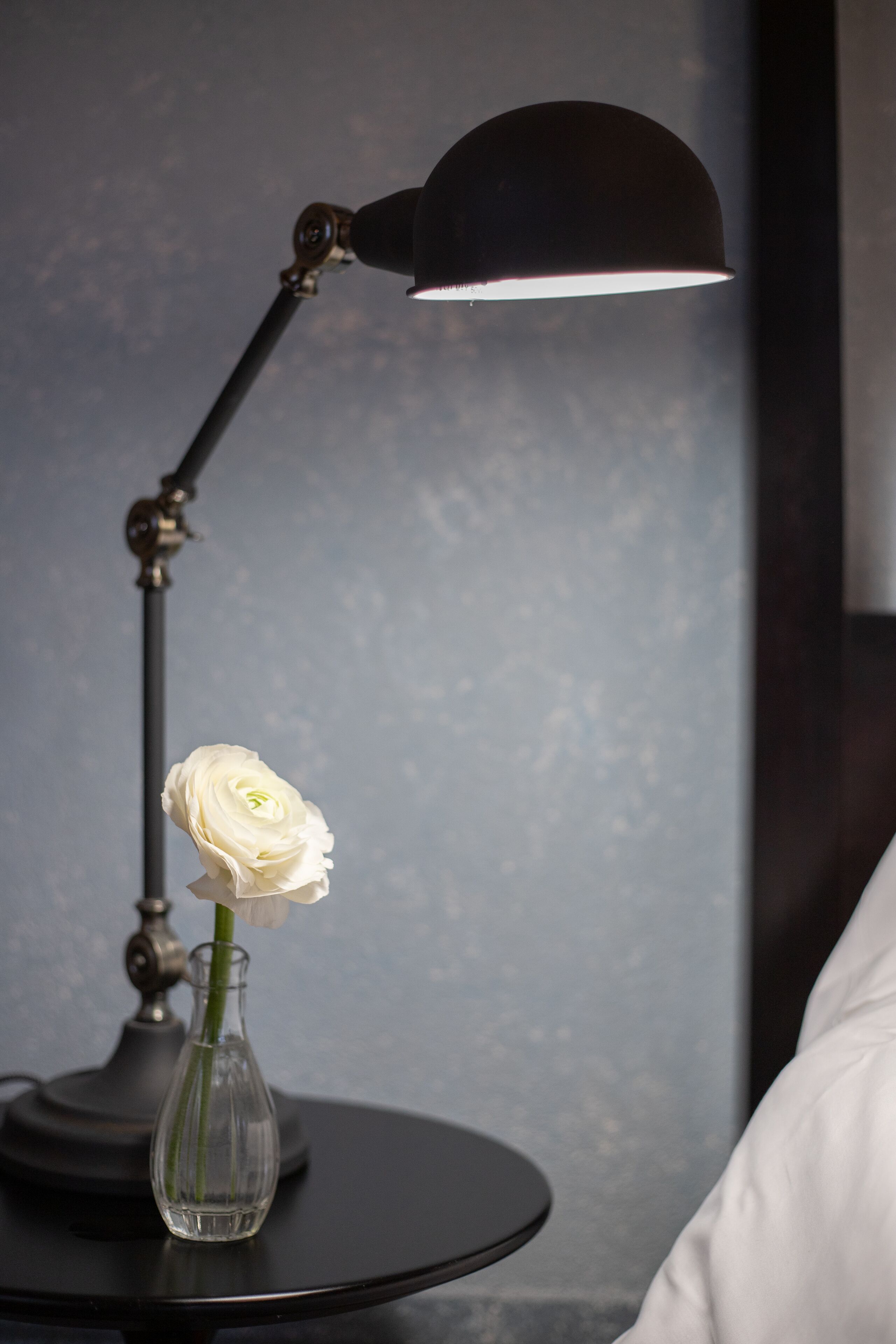 Een witte roos in een glazen vaas op een rond zwart bureau, met een zwarte bureklamp ernaast.