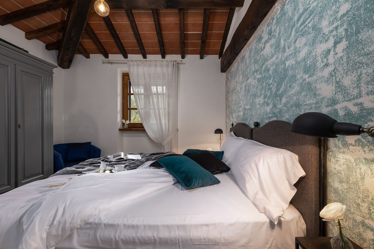 Un lit bien fait, avec des oreillers blancs et turquoise, une lampe de chevet et une fenêtre ornée de rideaux légers.