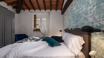Un letto rifatto con cuscini bianchi e color acquamarina, una lampada da comodino e una finestra con tende leggere.