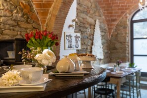 Un coin repas rustique avec un mur en pierre, une table en bois dressée avec des tasses blanches, une théière et un présentoir à gâteaux.
