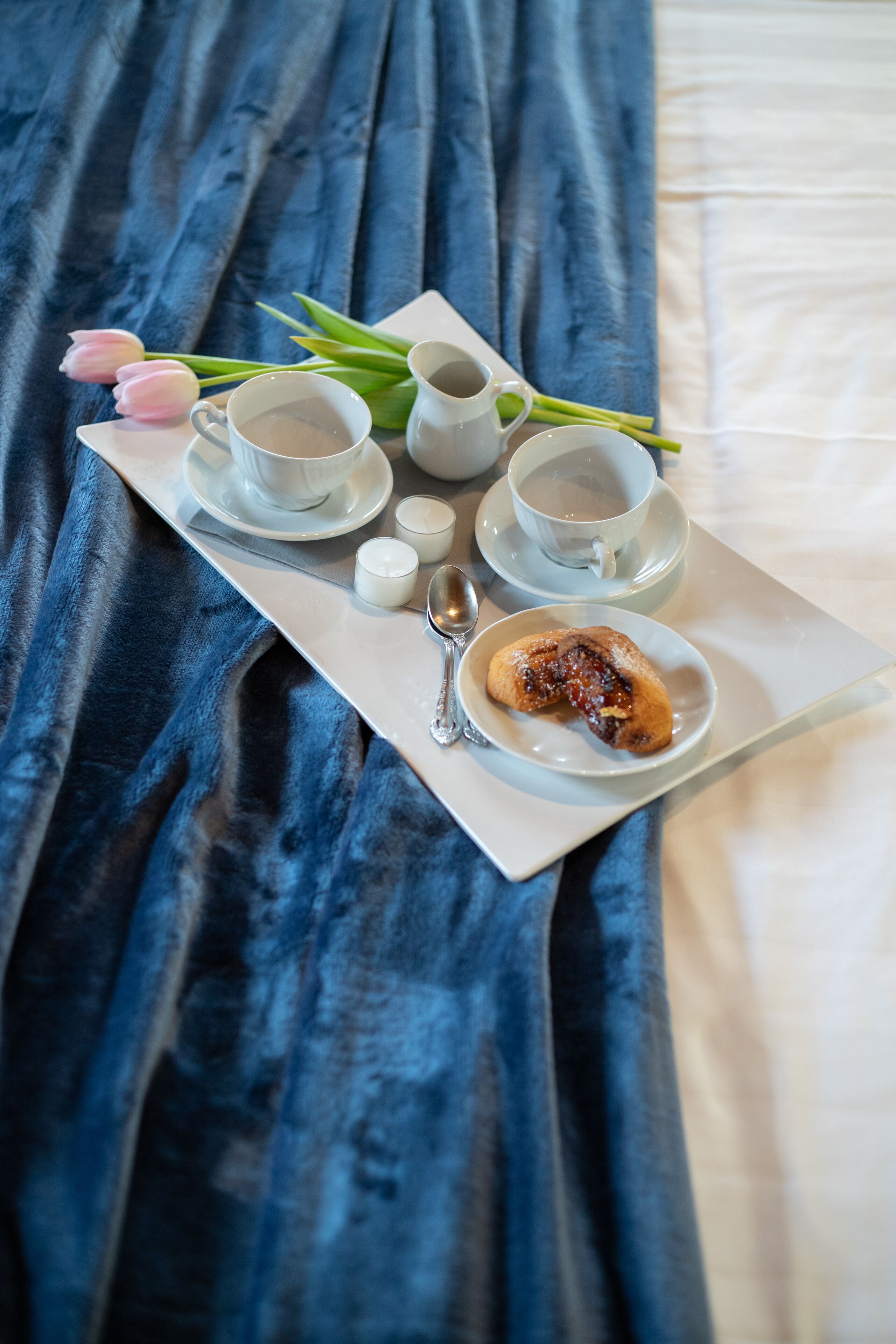 A tray with a cup of tea, a small bowl of jam, a spoon, and a tulip on a bed with a blue blanket.