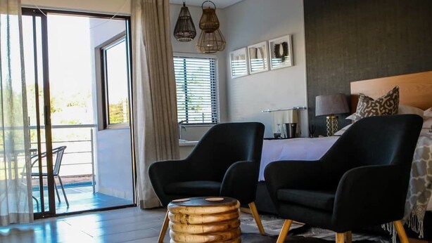 A modern bedroom with a bed, two black armchairs, a wooden side table, and a view of the outdoors through a sliding glass door. - eMfuleni Lifestyle (Lobamba)