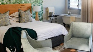 A hotel room with a bed, two armchairs, a small table, a bathroom with a bathtub, and a window with curtains. - eMfuleni Lifestyle (Lobamba)