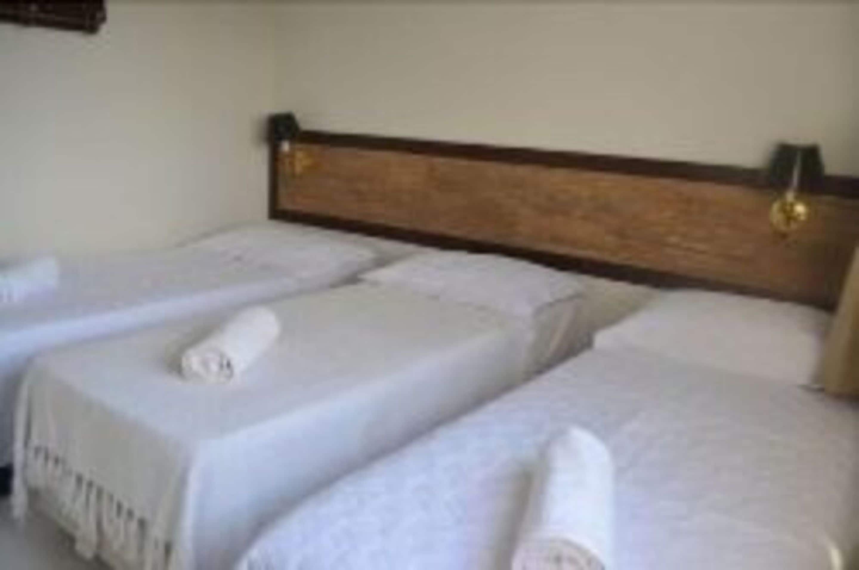 Habitación de hotel con dos camas individuales, cada una con una colcha blanca y una toalla enrollada.