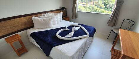 Cama bem arrumada com lençóis brancos e um cobertor azul, mesa de cabeceira de madeira, uma pequena mesa de madeira e vista para o verde pela janela.