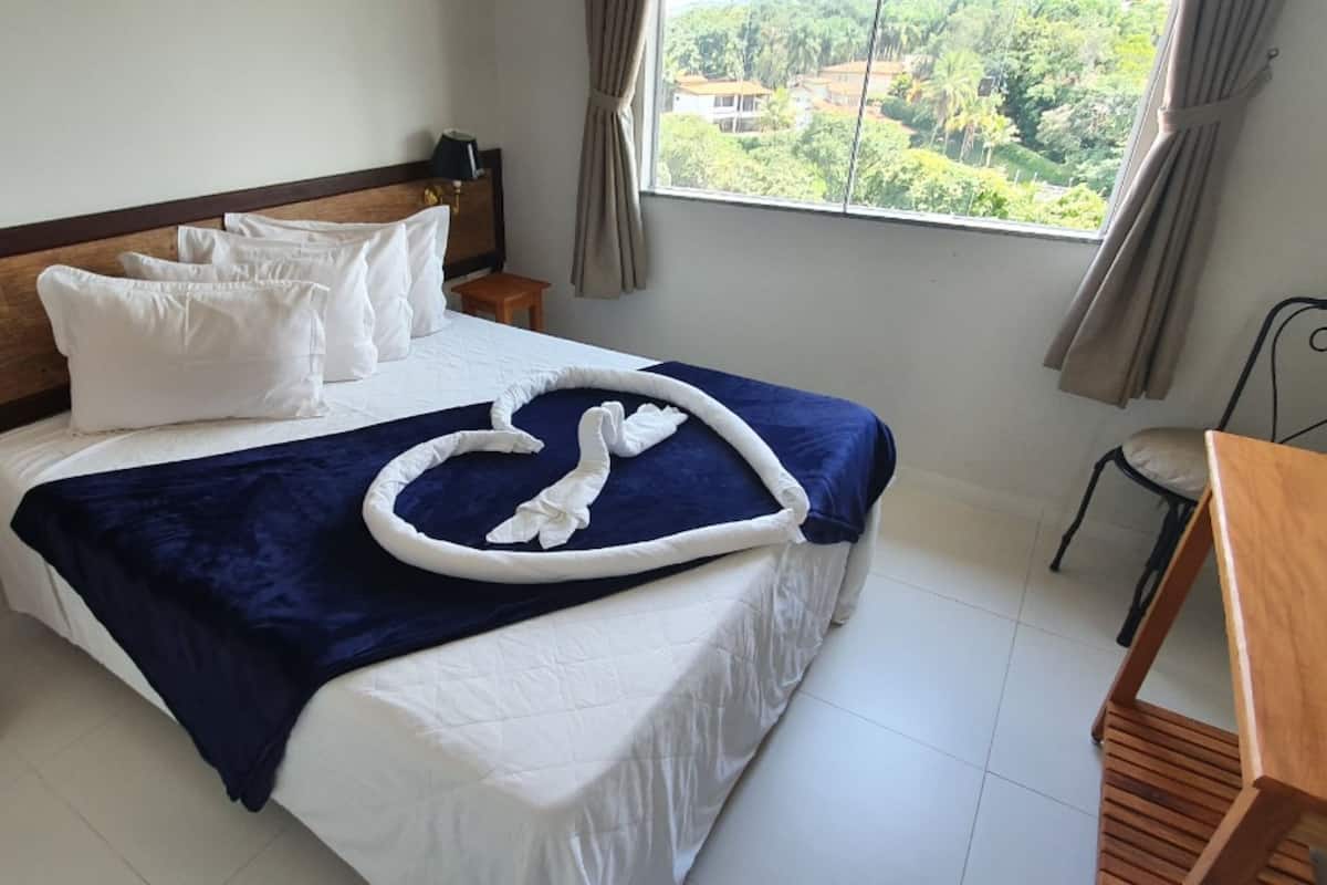 A hotel room with a bed, a wooden nightstand, a chair, a table, and a window with a view of greenery.
