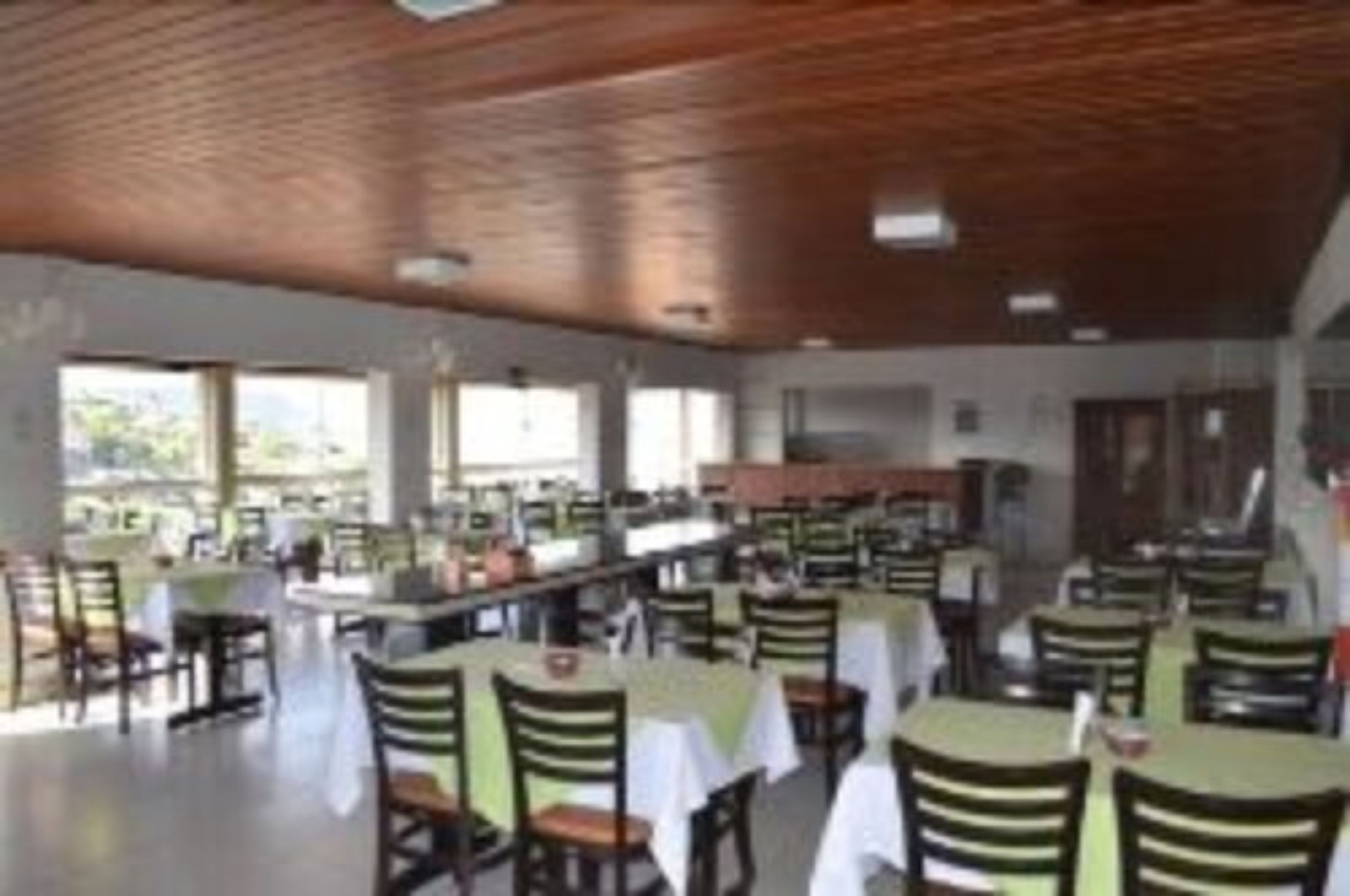 Un comedor amplio con varias mesas y sillas, ventanas grandes que dejan entrar luz natural y un techo alto con vigas de madera a la vista.