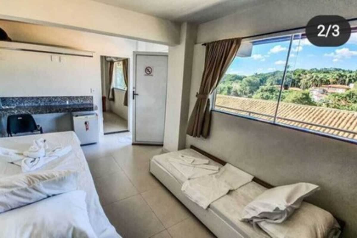 A hotel room with a bed, a sofa, a small kitchen area, and a view of greenery.