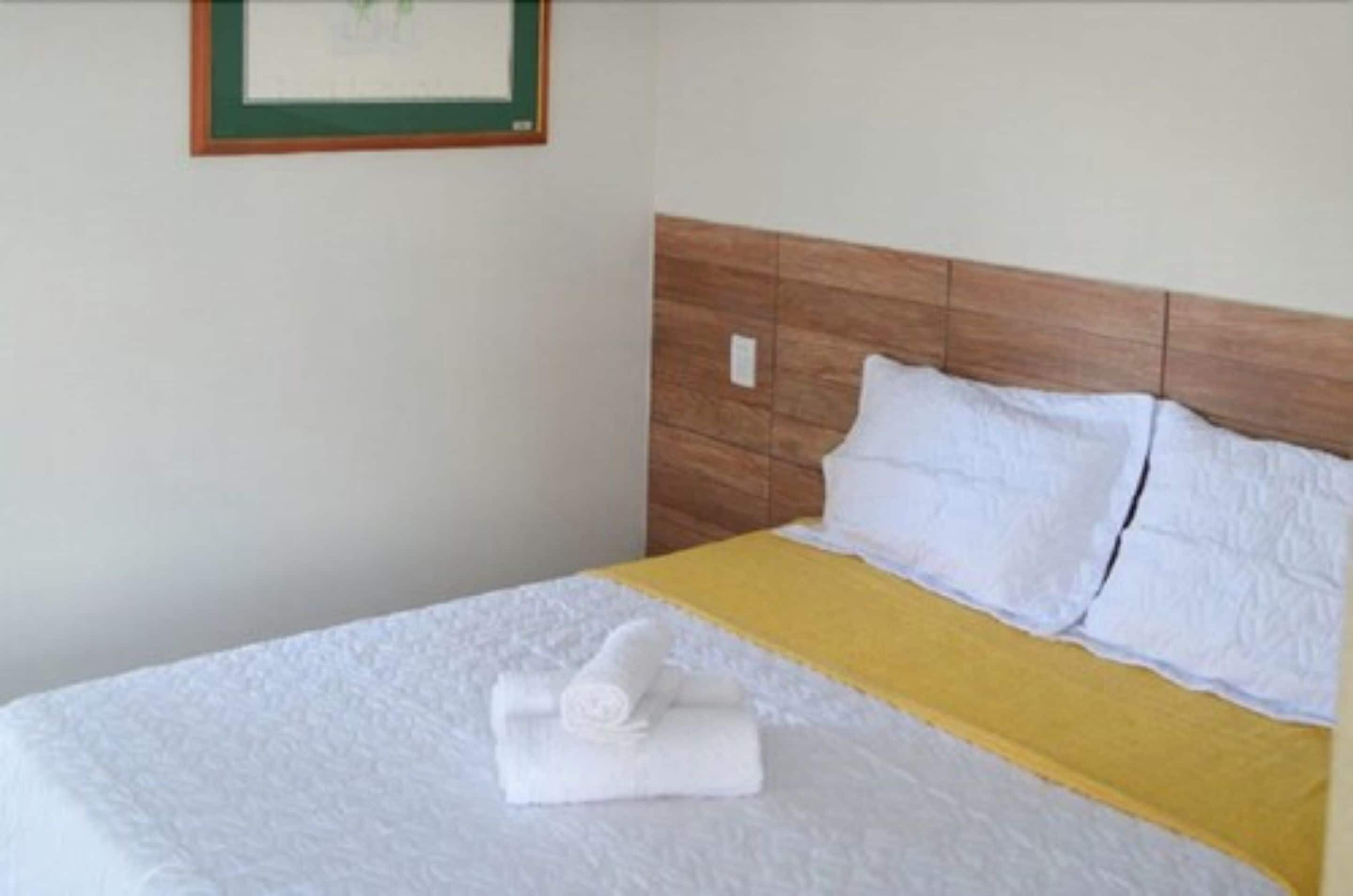 A neatly made bed with white linens and a yellow blanket, a wooden headboard, and a framed picture on the wall.