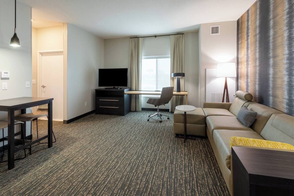 A hotel room with a desk, TV, sofa, and a large window with curtains.