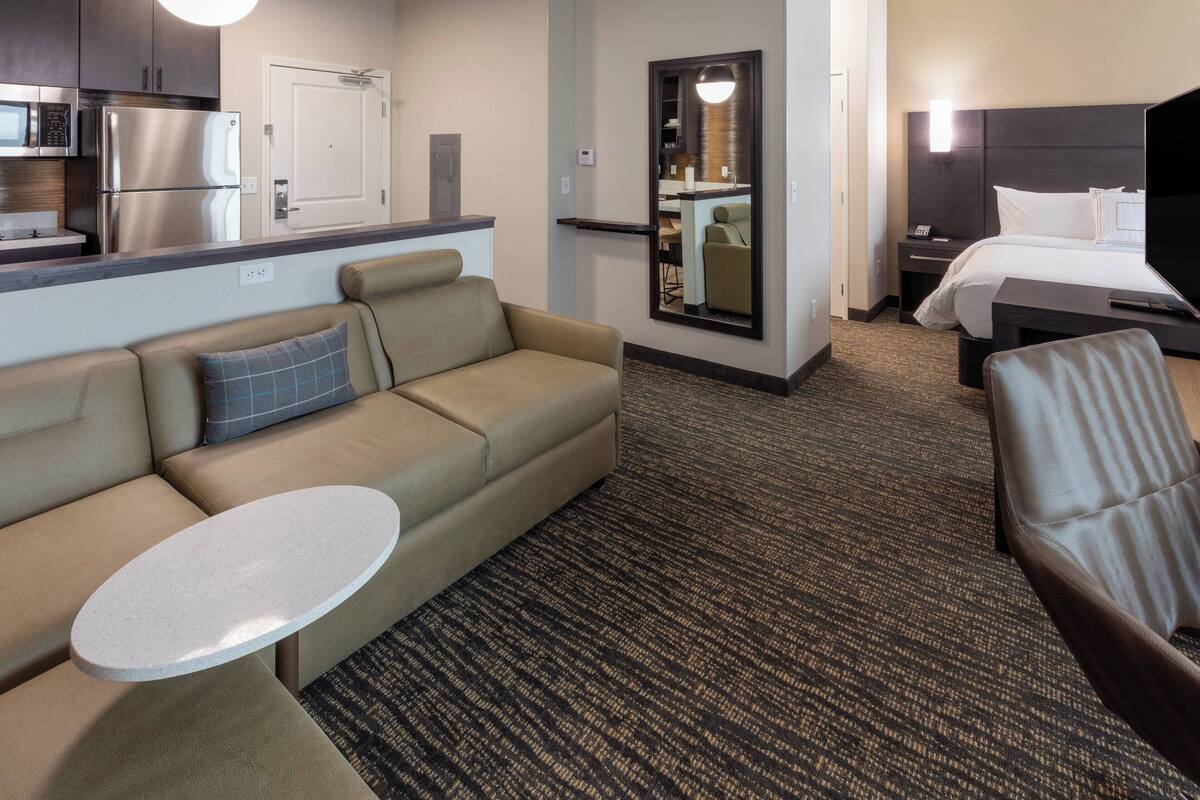 A hotel room with a sofa, a small round table, a bed, a nightstand, a TV, and a kitchen area with a refrigerator and microwave.