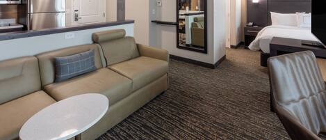A hotel room with a sofa, a small round table, a bed, a nightstand, a TV, and a kitchen area with a refrigerator and microwave.