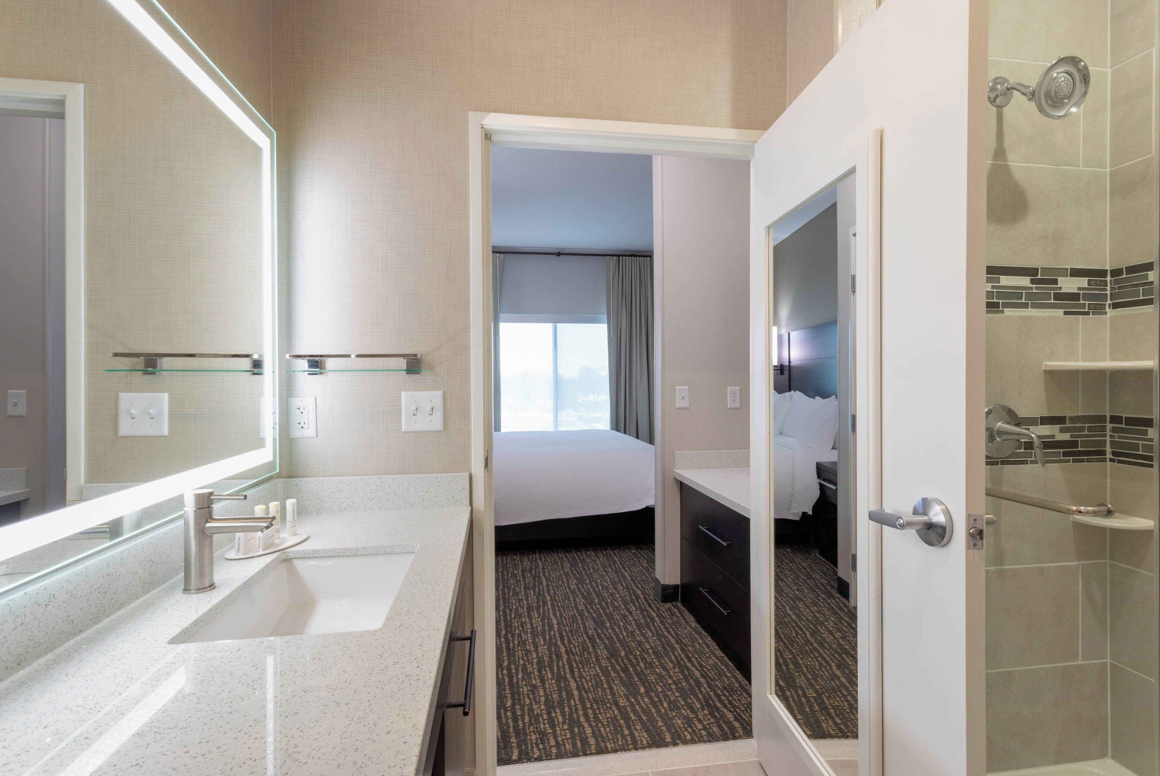 A modern hotel bathroom with a large mirror, a sink, a shower, and a bed visible through an open door.