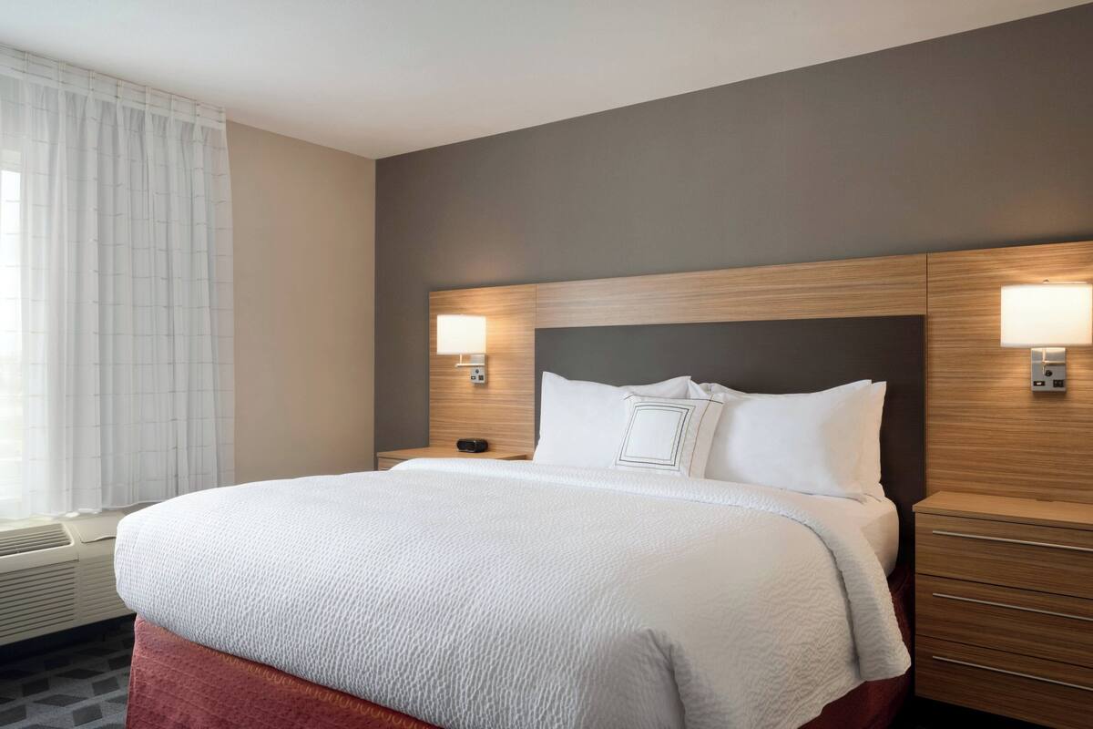 A hotel room with a large bed, two bedside tables with lamps, a window with curtains, and a wall-mounted air conditioning unit.