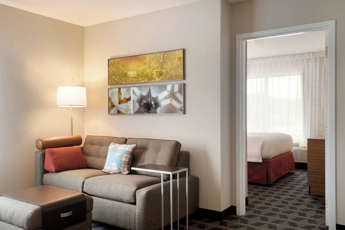 A hotel room with a sofa, a small table, a lamp, and a bed with a red bedspread. There is a painting on the wall and a window with curtains.