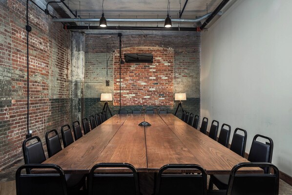 A conference room with a long wooden table, black chairs, two wall-mounted lamps, and exposed brick walls.