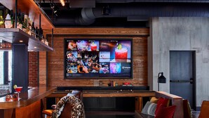 A bar area with a wooden bar counter, a television displaying various content, and seating with cushions. - Moxy Chattanooga Downtown (Chattanooga)