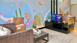 A living room with a wall mural of an underwater scene, a television, a gaming console, and a small table with a plant.