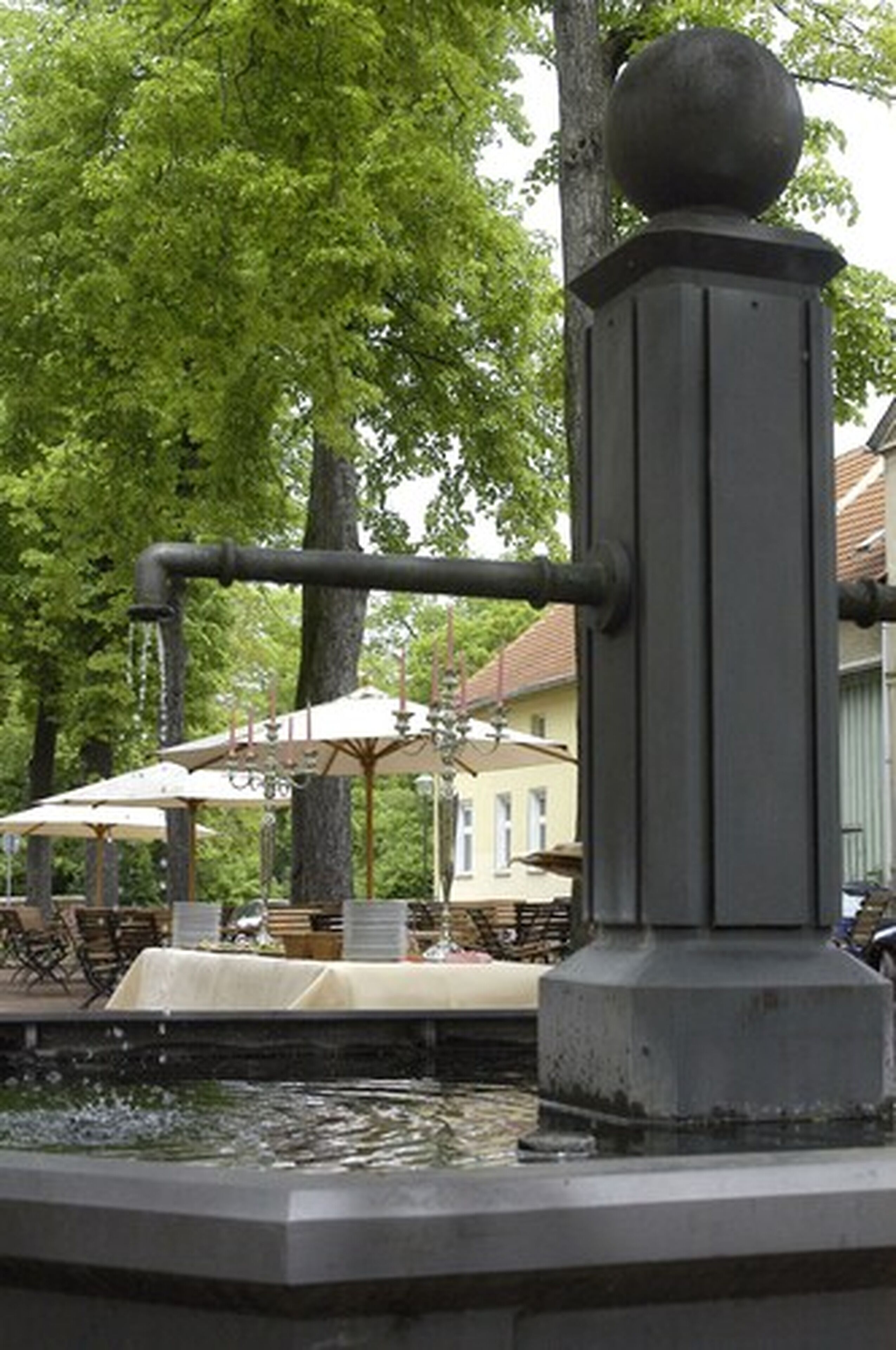 Ein Brunnen mit einer Wasserpumpe, umgeben von Bäumen und Sitzgelegenheiten mit Sonnenschirmen.
