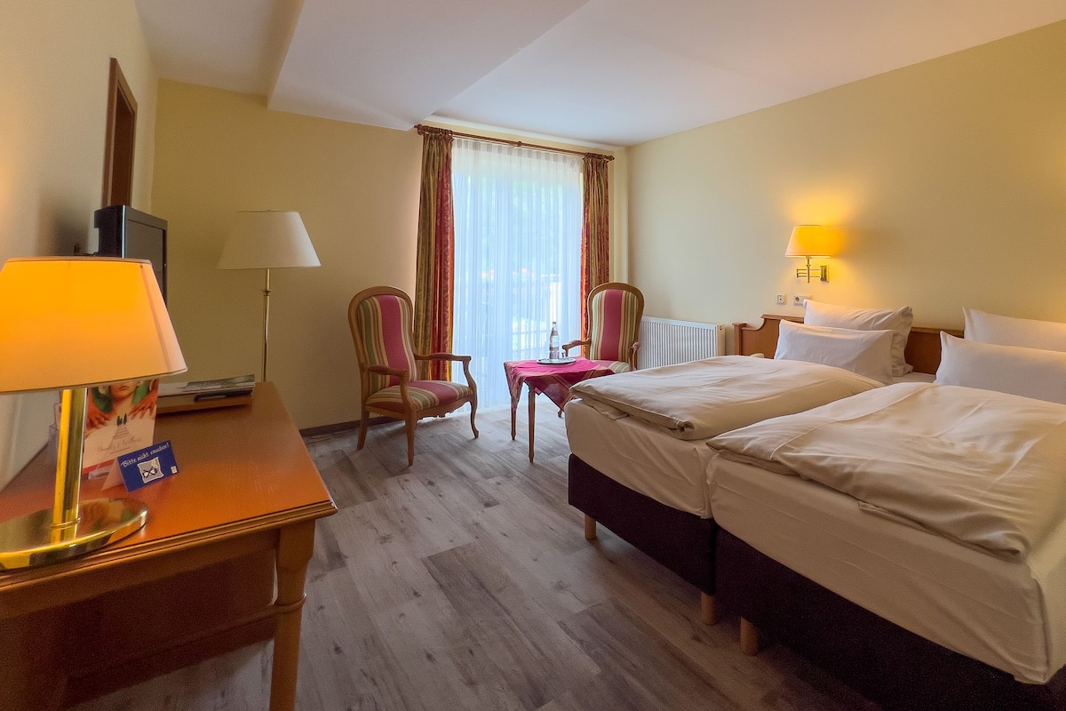 A hotel room with two beds, a desk with a lamp, a chair, and a window with curtains.