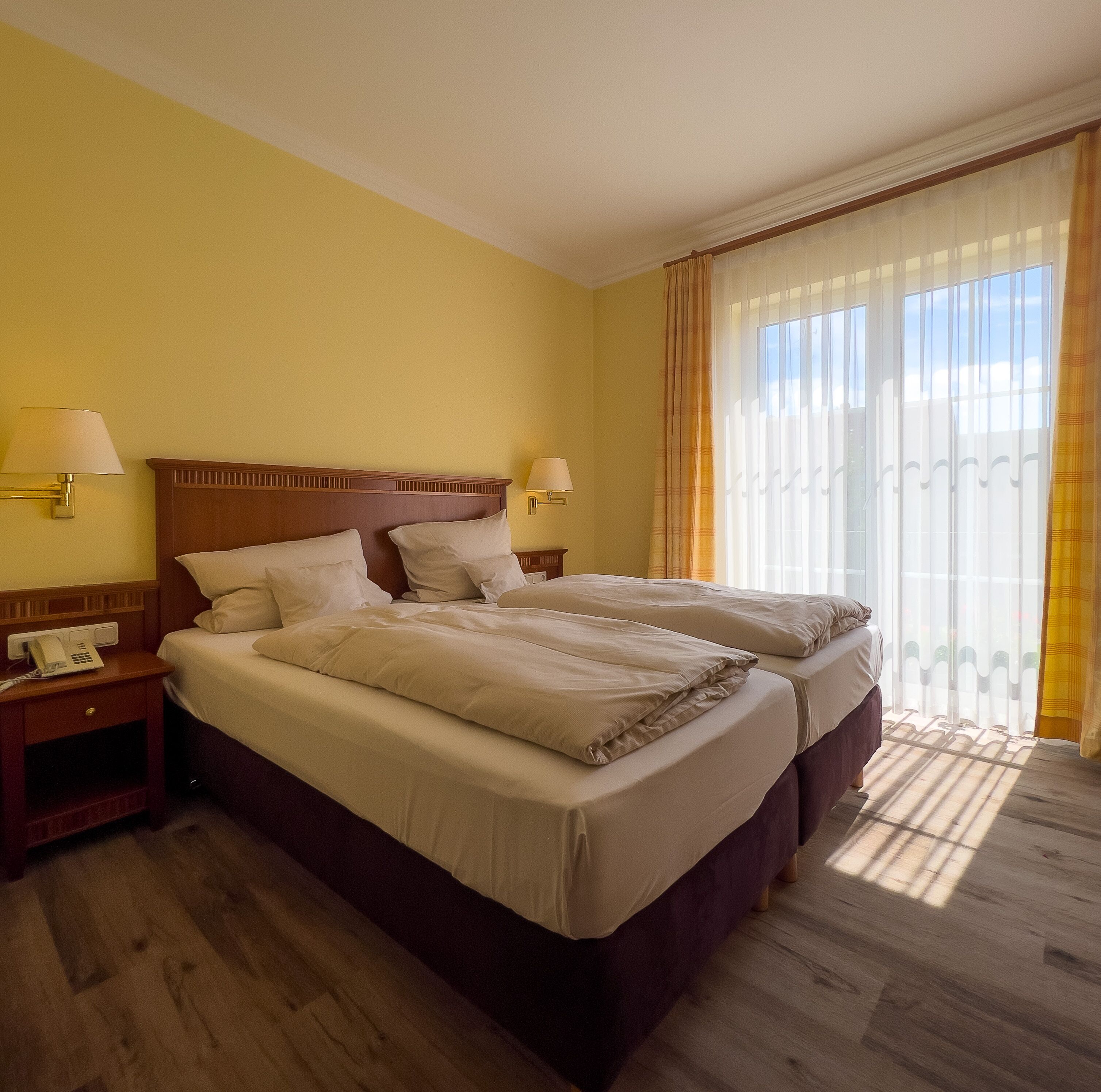 Ein Hotelzimmer mit zwei Betten, Holz-Kopfteilen, Nachttischlampen, einem Nachttisch mit Telefon und einem großen Fenster mit durchsichtigen Vorhängen.