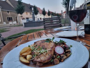 A meal with a steak, potatoes, and mushrooms on a white plate, accompanied by a glass of red wine, set on an outdoor wooden table.
