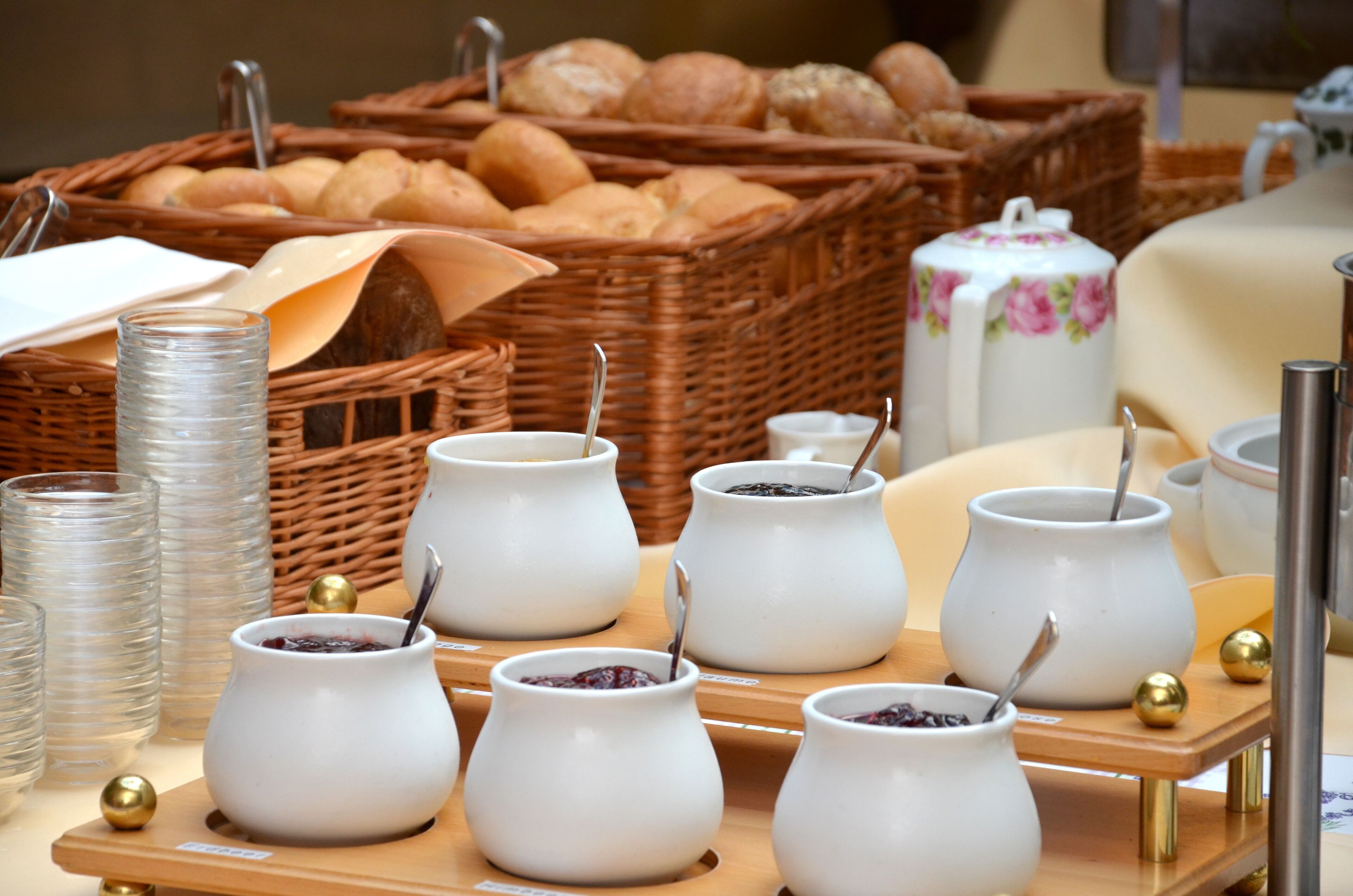 A buffet setup with white ceramic dishes containing food, a wicker basket with bread, and a floral-patterned teapot.
