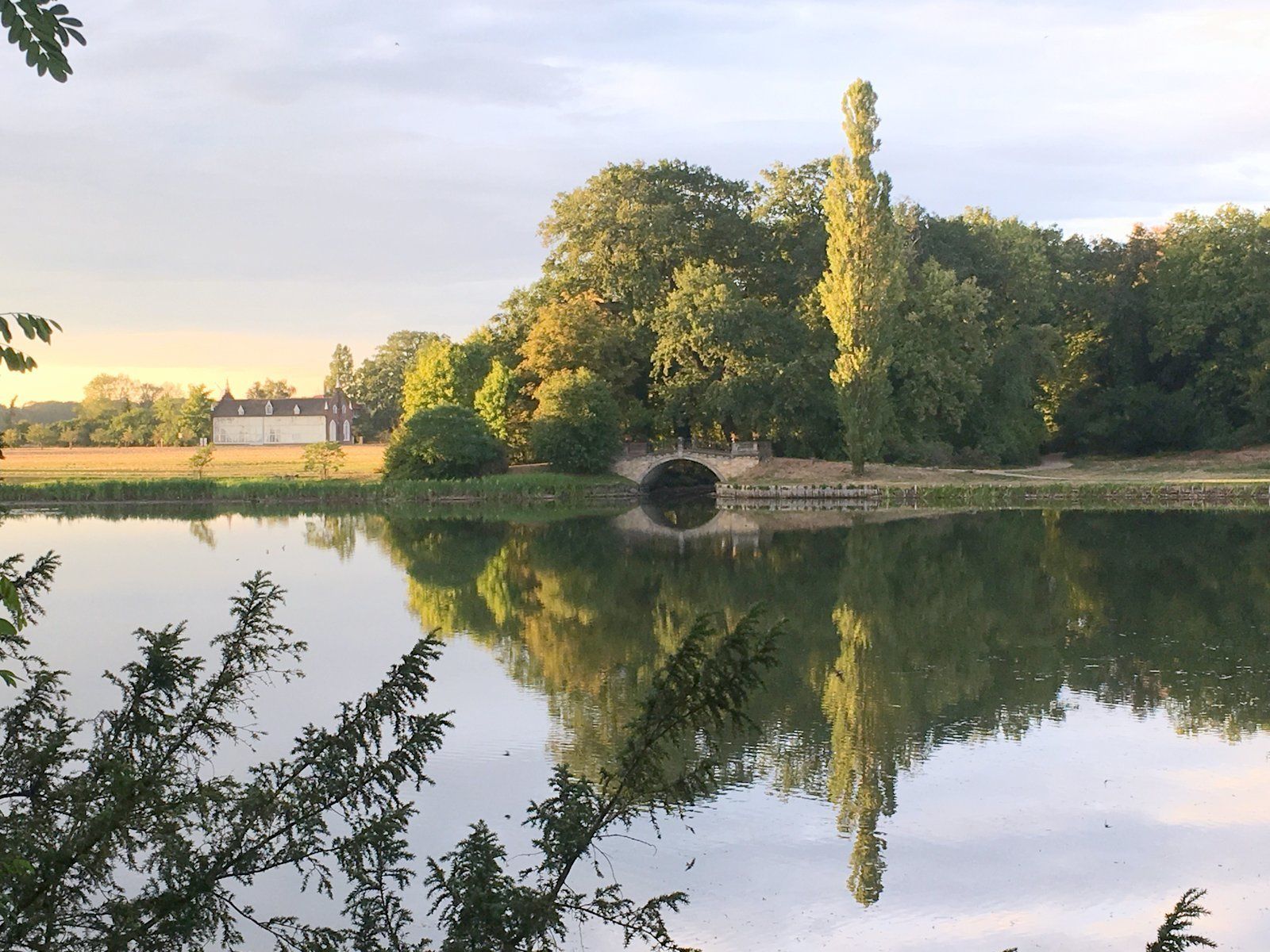 Eine friedliche Landschaft mit einer Brücke über ruhigem Wasser, umgeben von üppigem Grün und einem Gebäude in der Ferne.