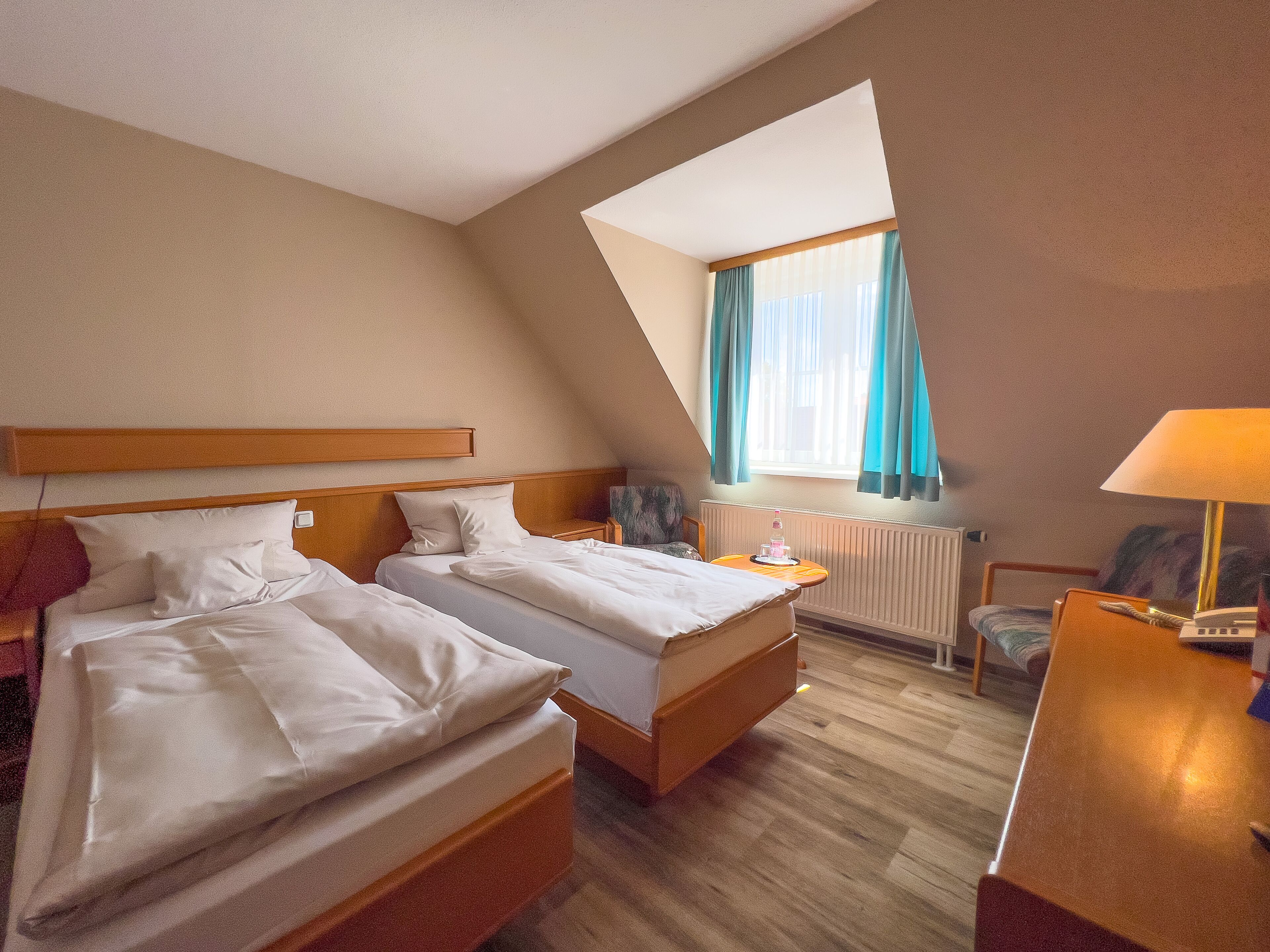 A hotel room with two beds, a small table, a chair, a lamp, and a window with curtains.