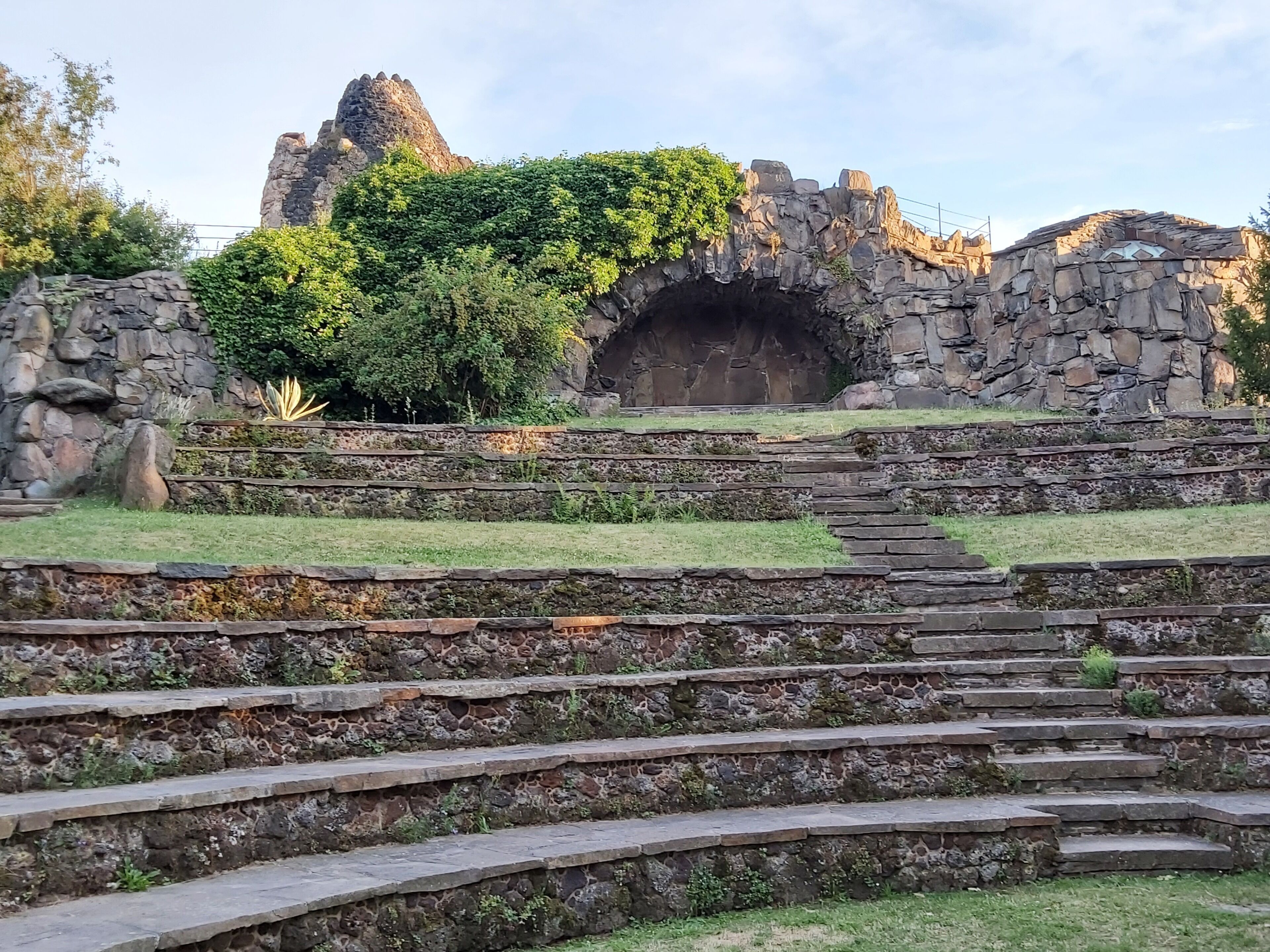 Una escalera de piedra que conduce a la entrada de una cueva, rodeada de follaje y una estructura rocosa.
