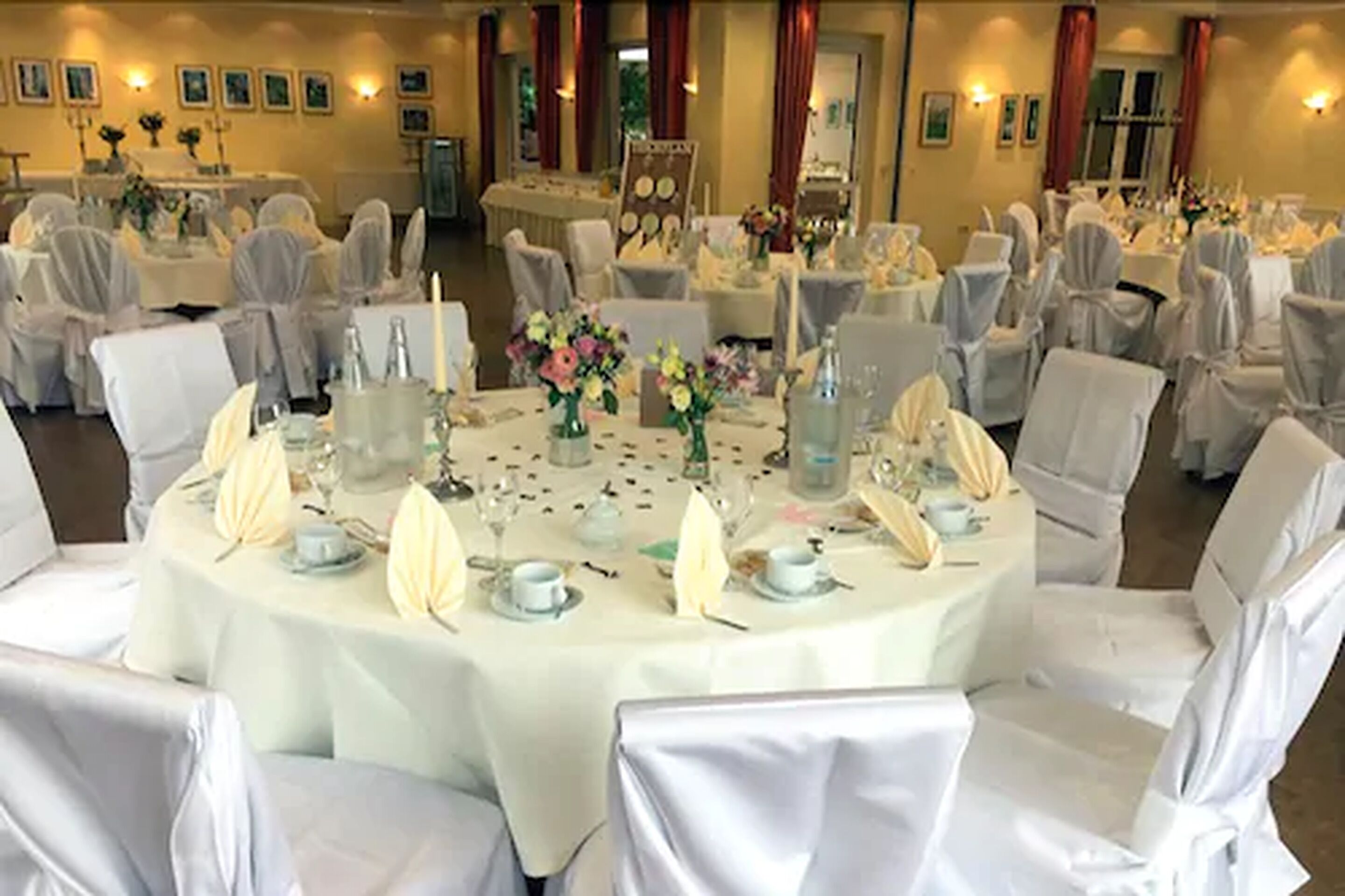 A banquet hall with round tables set for a formal event, each with white tablecloths, chairs with white covers, and floral centerpieces.