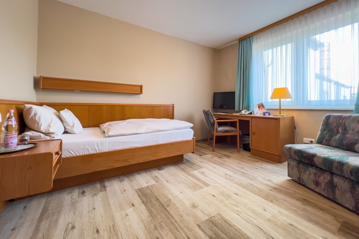 A hotel room with a bed, a desk, a chair, a sofa, a TV, and a window with curtains.