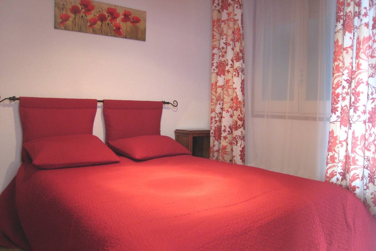 A bed with red bedding, a wooden nightstand, a painting of flowers on the wall, and floral curtains.