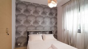 A neatly made bed with white linens, a headboard with a geometric pattern, and a window with sheer curtains. - White Rose Villa (Kassandra)