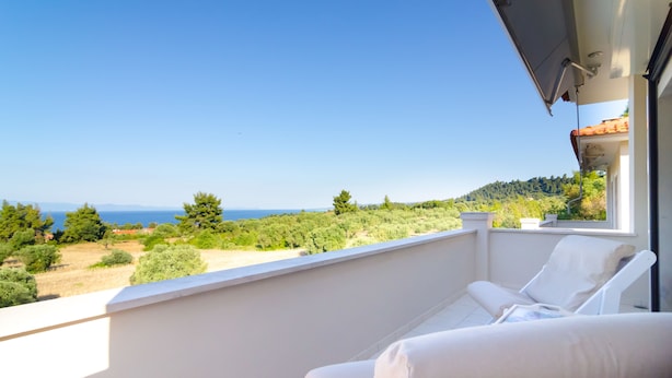 A balcony with a white cushioned chair and a small table, overlooking a scenic view of the sea and greenery. - White Rose Villa (Kassandra)