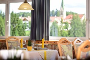 A dining table set with glasses, a candle, and a potted plant, with a view of houses and trees through large windows. - Hotel Tirolerhof (Terento)