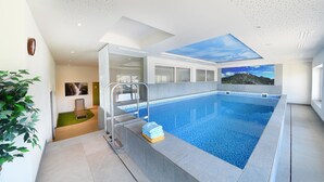 A modern indoor swimming pool with a skylight, a glass door leading to a room, and a mountain view mural. - Hotel Tirolerhof (Terento)