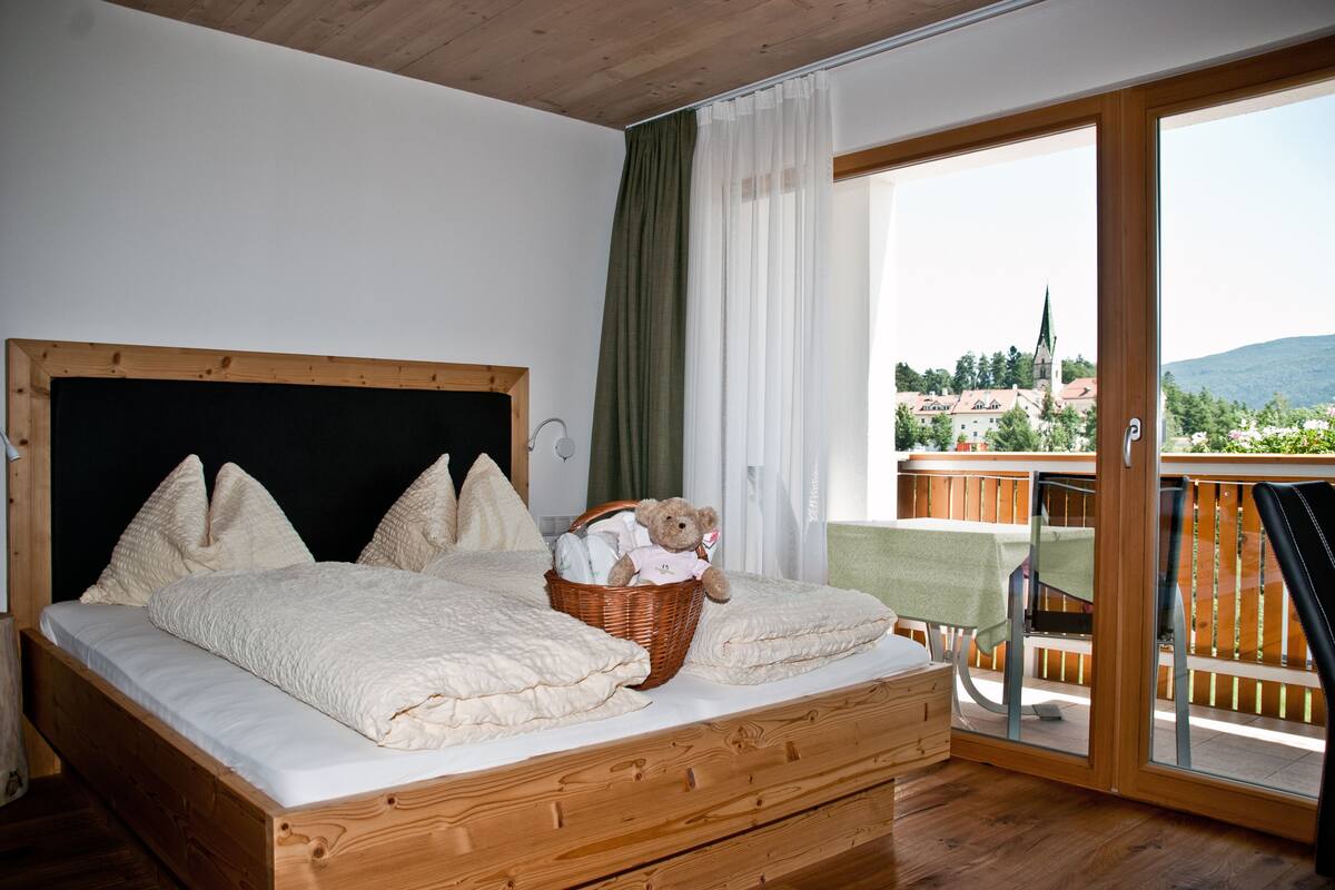 Une chambre avec un lit en bois, un panier en osier contenant un ours en peluche, et un balcon avec une table et des chaises.