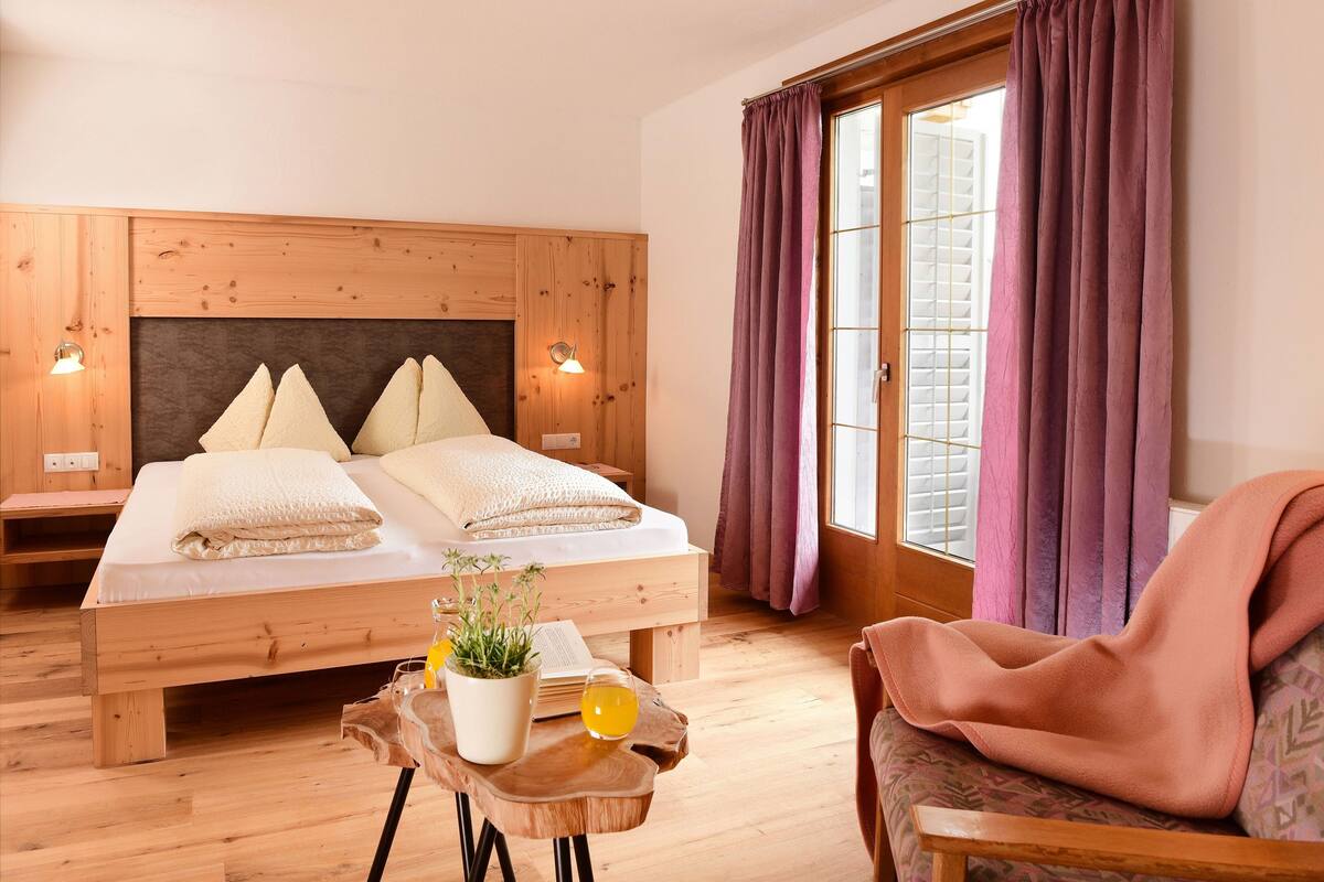 Un lit en bois avec deux oreillers, une table de chevet avec une plante et un verre de jus d’orange, et une chaise avec une couverture.