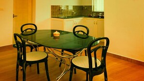 A dining area with a glass table and four chairs, a small table with a decorative item, and a kitchen in the background. - Magic Land Resort (Chalakudy)