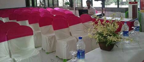 A conference room with rows of chairs covered in white and pink fabric, a table with water bottles and a potted plant, and a person at a laptop in the background.