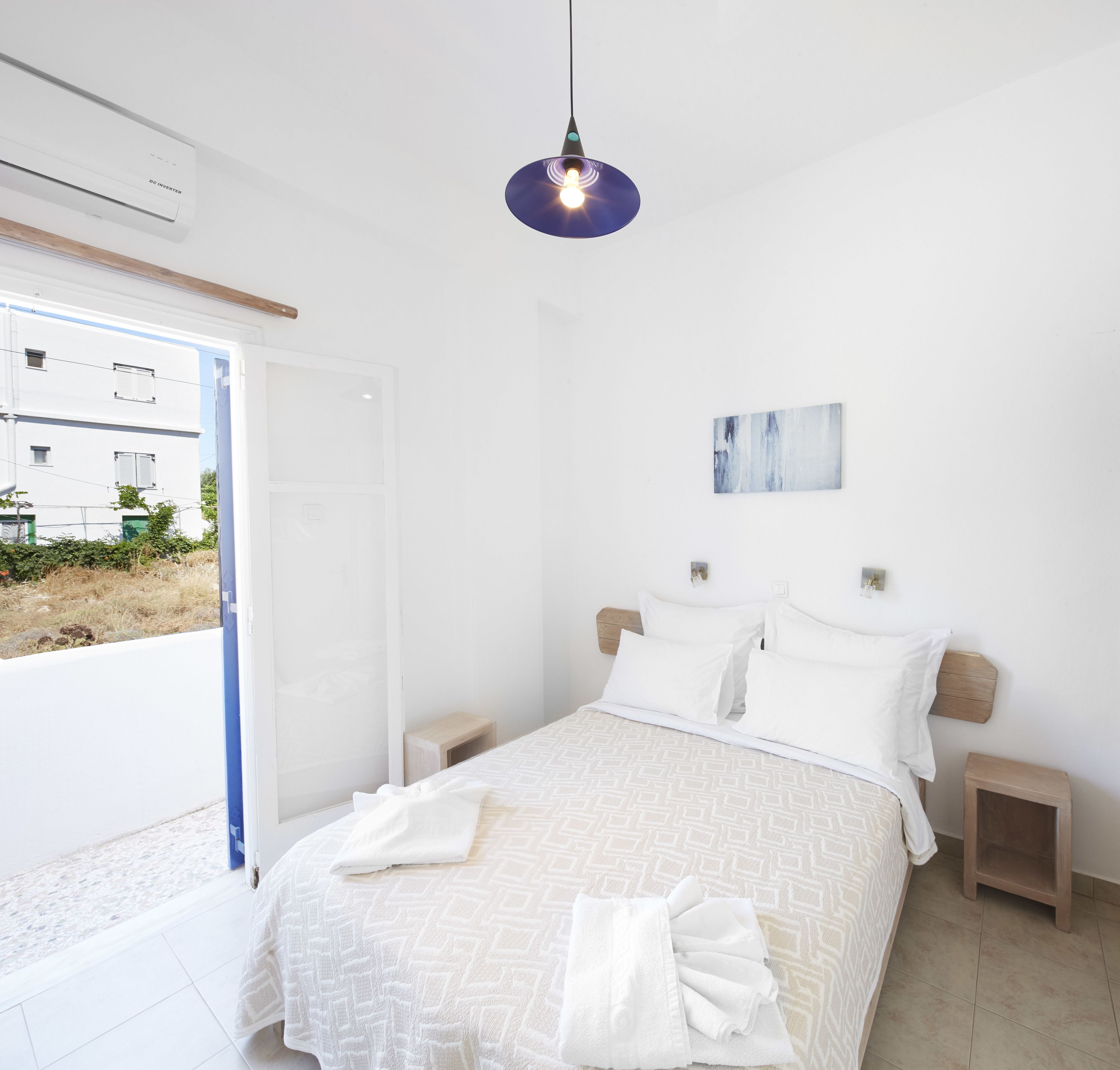 A bedroom with a bed, bedside tables, a hanging light fixture, a door, and a view of buildings outside.