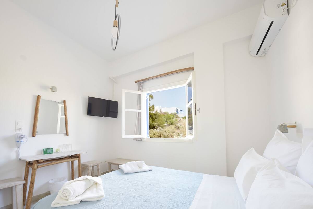 Un dormitorio con cama, mesita de noche, espejo, ventana con contraventanas abiertas y televisor montado en la pared.